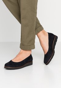 Bailarinas de ante negro con diseño de punta redonda, tacón bajo en cuña y suela texturizada, combinadas con pantalones enrollados de color verde oliva.