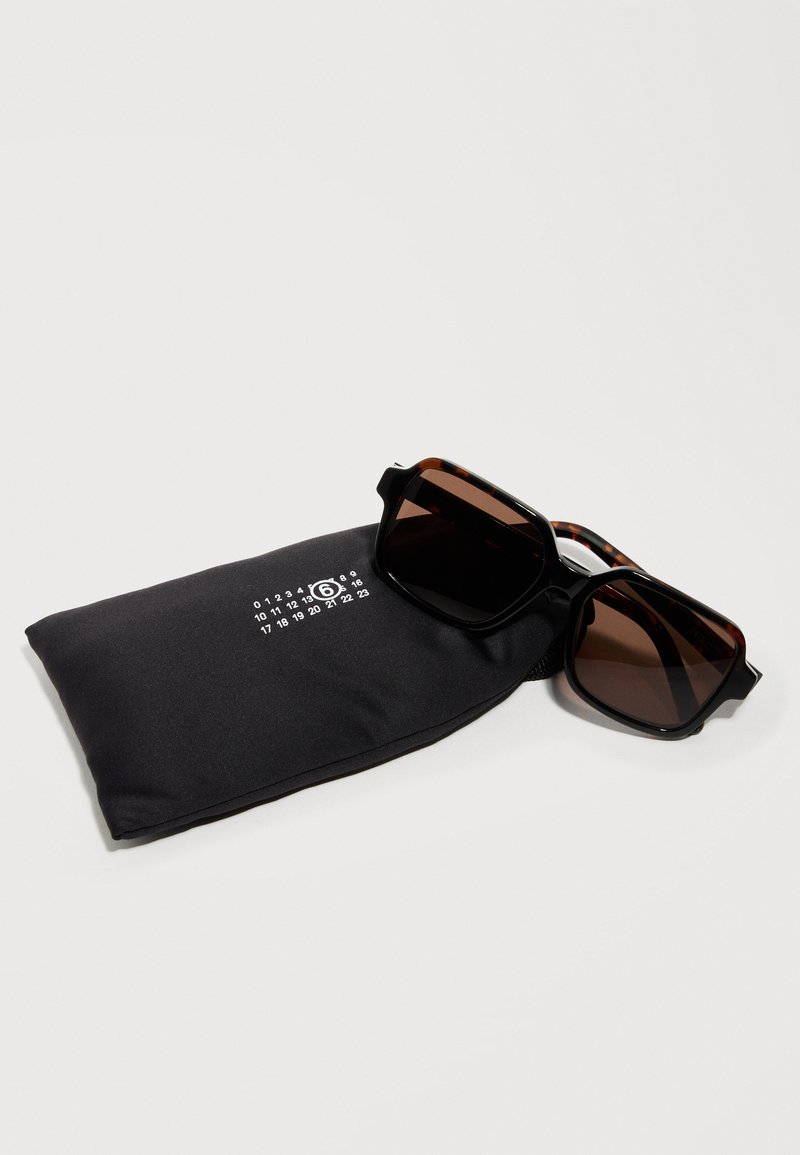 Schwarze rechteckige Sonnenbrille mit braunen Gläsern, die auf einem schwarzen Stoffbeutel mit weißer Zahlenaufdruck auf einer weißen Oberfläche liegt.