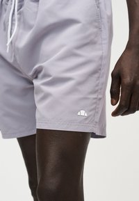 Κοντινό πλάνο ενός ατόμου που φορά ανοιχτό γκρι σορτς με λευκά κορδόνια και μικρό λευκό λογότυπο κοντά στο τελείωμα στο αριστερό μηρό.