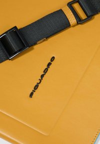 Borsa in pelle gialla con tracolla regolabile nera e logo "Piquadro" impresso in rilievo sulla patta frontale nera.