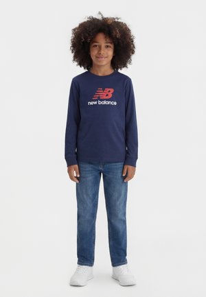 Bambino con capelli ricci che indossa una maglia a maniche lunghe blu navy di New Balance, jeans blu e scarpe da ginnastica bianche, in piedi davanti a uno sfondo bianco neutro.