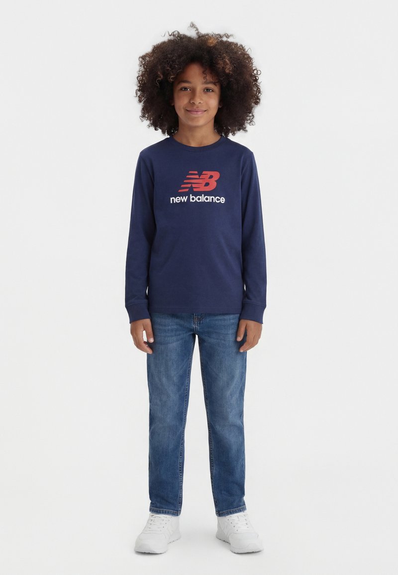 Kind mit lockigem Haar, das ein marineblaues New Balance Langarmshirt, eine blaue Jeans und weiße Turnschuhe trägt und vor einem schlichten weißen Hintergrund steht.
