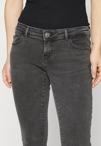 Donna che indossa jeans skinny neri sbiaditi a vita alta e un top a coste nero, con le mani rilassate ai lati, su uno sfondo neutro.
