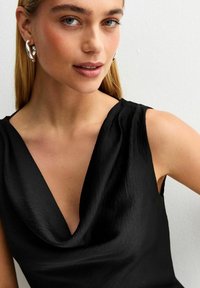 Top senza maniche nero con scollo drappeggiato e texture liscia. La modella indossa orecchini a cerchio grandi in argento. Lo sfondo è semplice.