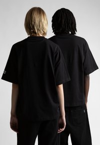 Čierne bavlnené tričká s krátkymi rukávmi, vo voľnom strihu, so štítkami, nosené dvomi osobami zozadu na neutrálnom pozadí.