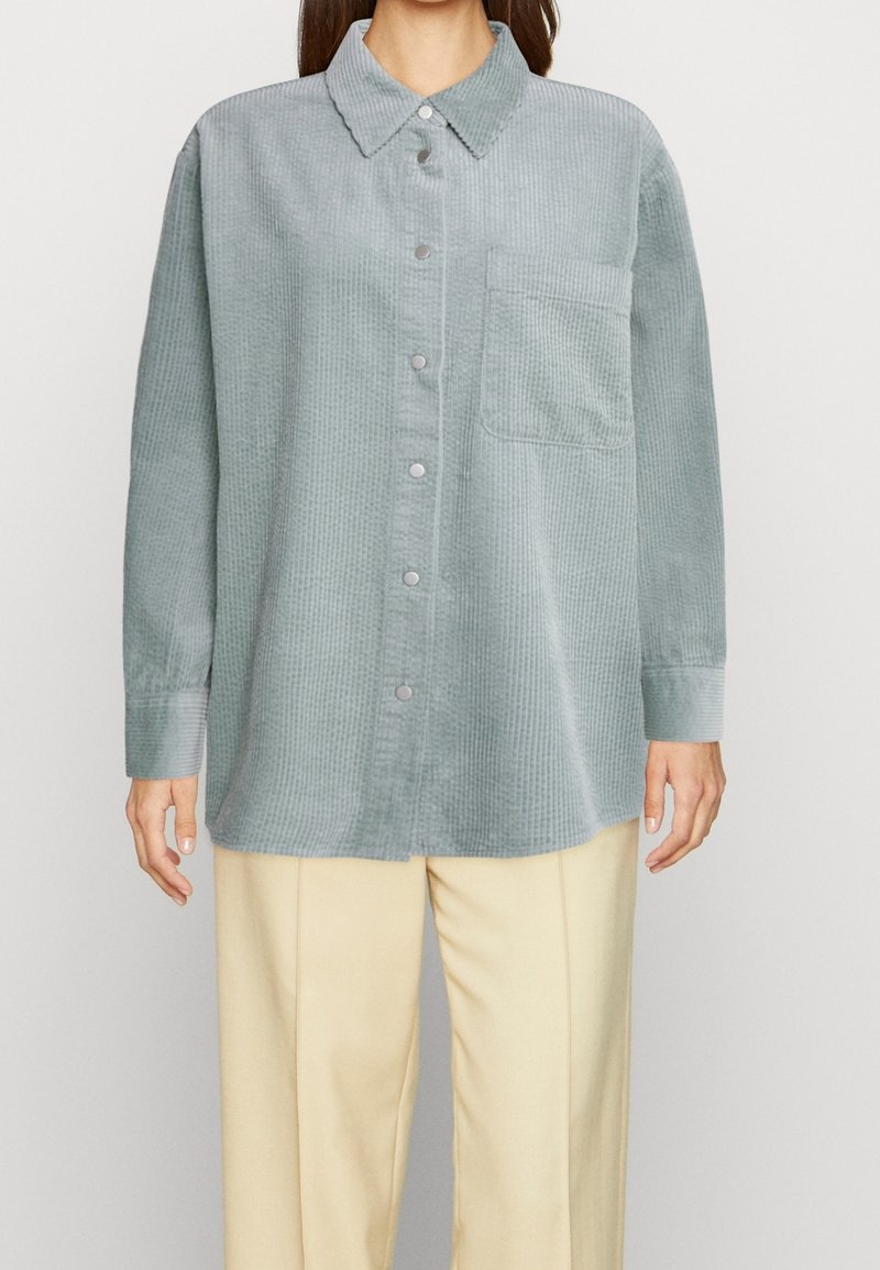 Femme portant une chemise ample en velours côtelé bleu-gris pâle avec une poche poitrine unique et un pantalon couleur crème.