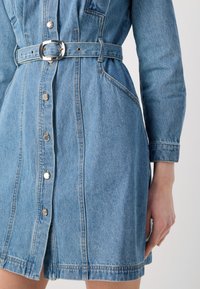 Lys blå denimkjole med lange ermer, knappestolpe foran, innsnevret midje med et sølvbelte og sidelommer. Glatt tekstur og uformelt design.