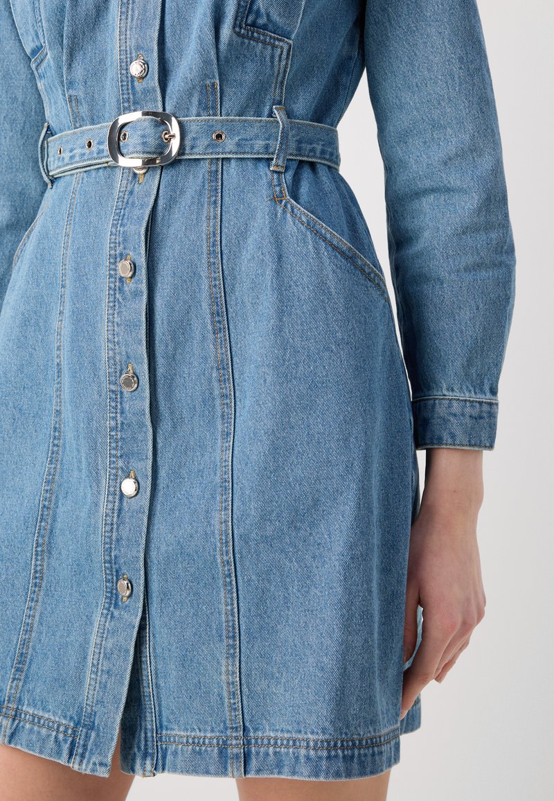 Robe en denim bleu clair avec des manches longues, un boutonnage sur le devant, une taille cintrée avec une ceinture en argent, et des poches latérales. Texture lisse et design décontracté.