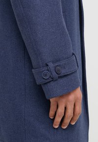 Prato ravvicinato di un cappotto in lana blu navy, caratterizzato da un tessuto texturizzato, un polsino con due bottoni scuri e una tasca laterale.