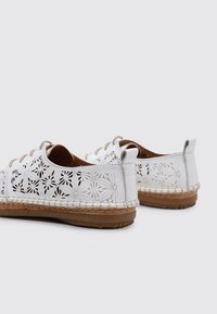 Zapatos de piel blanca perforada con patrones florales, suelas de goma marrón y diseño de cordones. Los acentos incluyen detalles trenzados a lo largo de la suela.