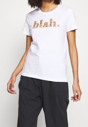 Persoon draagt wit shirt met korte mouwen met gouden glittertekst "blah.", gecombineerd met losse zwarte broek tegen een effen achtergrond.