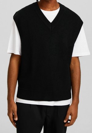 Ein Mann trägt eine schwarze ärmellose V-Ausschnitt-Strickweste über einem weißen kurzärmligen T-Shirt mit schwarzen Hosen vor einem schlichten Hintergrund.