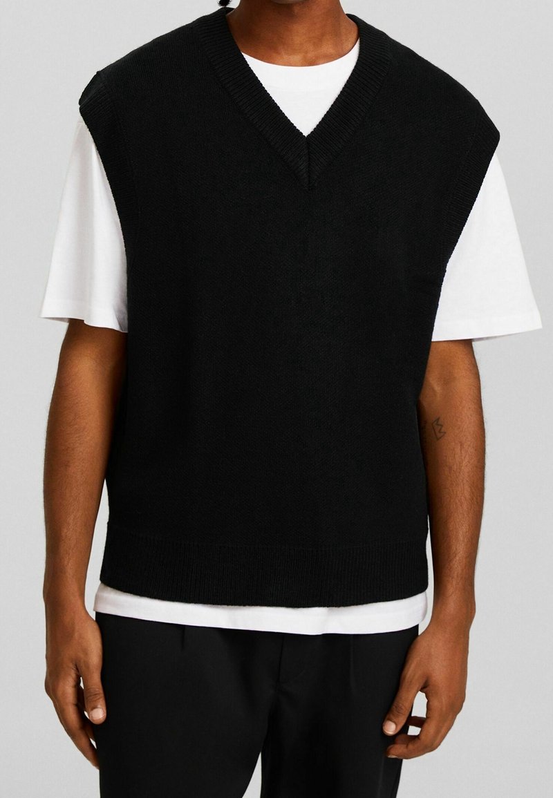 Homme portant un gilet en maille noir sans manches à col en V par-dessus un t-shirt blanc à manches courtes avec un pantalon noir sur un fond uni.