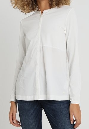 Vrouw draagt een wit overhemd met lange mouwen en knopen, gecombineerd met donkerblauwe jeans, staand tegen een effen lichte achtergrond.