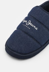 Zapatilla de color azul marino fabricada con material similar al denim, con acolchado, punta redondeada y logo de la marca: "Pepe Jeans London" en blanco.
