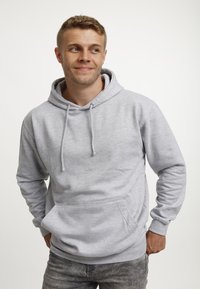 Uomo giovane che sorride, indossando una felpa grigia e jeans sbiaditi, in piedi con le mani nelle tasche, mentre guarda di lato contro uno sfondo semplice.