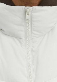 White padded jacket with a high collar and visible zipper, features textured fabric and contrasting brown interior lining.
