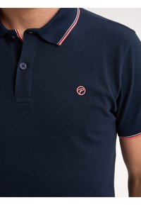 Námořnická modrá polo tričko vyrobené z bavlny, se solidním vzorem, s tří knoflíkovým zapínáním a s kontrastními červenými a bílými akcenty na límci.