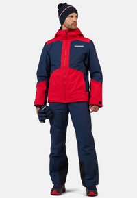 Giacca da sci uomo rossa e blu navy con zip frontale, cappuccio e dettagli neri. Abbinata a pantaloni blu navy e guanti blu navy. Tessuto texturizzato.