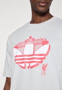 Ανοιχτό γκρι βαμβακερό t-shirt με μεγάλη κόκκινη γραφική παράσταση του σήματος της Liverpool FC και το εμβληματικό κείμενο "Δεν θα είστε ποτέ μόνος" από �πάνω.