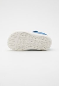 Blaues Lederschuh mit einer weißen Gummisohle, ausgestattet mit kreisförmigen Grip-Mustern und einem Riemen zum Schließen. Texturierte Oberfläche mit gepolsterten Akzenten.