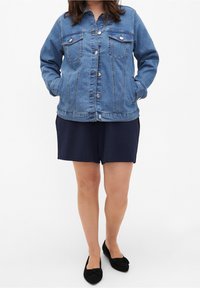 Ljusblå denimjacka med knapplås framtill, två bröstfickor och en avslappnad passform. Bärs över marinblå shorts och svarta ballerinas.