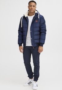 Marineblå pufferjakke med en fårskindsforet hætte, hvide snore, et mærke på brystet, parret med marineblå joggingbukser og hvide sneakers.