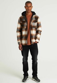 Chaqueta de franela en un patrón de cuadros marrón, crema y azul con capucha negra. Combinada con una camiseta de color óxido y jeans oscuros. Zapatillas gris.