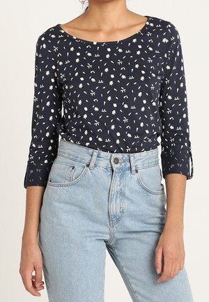 Granatowy top z długim rękawem w biały wzór kwiatowy, zestawiony z jasnoniebieskimi, wysokim stanem dżinsami, na stojącej osobie.