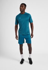 Sportig outfit bestående av en teal kortärmad skjorta och matchande shorts, tillverkade av andningsbart material, kombinerat med svarta sportskor och vita strumpor.