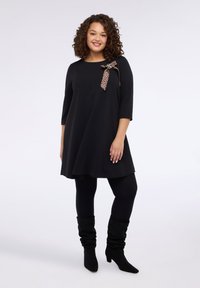 Czarna sukienka z rękawami do łokcia, ozdobiona kokardą w panterkę na ramieniu. Noszona z czarnymi legginsami i luźnymi czarnymi botkami.