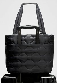 Crna torba s pravokutnim uzorkom, mekane teksture, dužih ručki, smještena iznad ručke crne kofer.