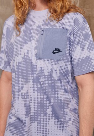 Personne portant un t-shirt Nike violet clair à motifs avec une poche poitrine zippée et un logo noir visible.