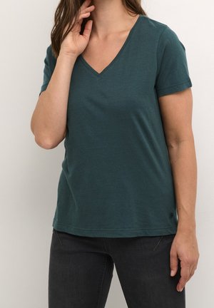 Kobieta w ciemnozielonej koszulce z krótkim rękawem i dekoltem w serek oraz czarnych spodniach, stojąca z jedną ręką przy twarzy, a drugą swobodnie opuszczoną wzdłuż ciała.