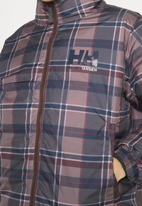 Gros plan d'une personne portant une veste zippée à carreaux marron et violet Helly Hansen, main dans la poche.