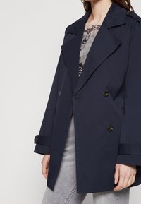 Granatowy, podwójnie zapięty trencz wykonany z gładkiego materiału, z wyraźnymi klapami, dużymi guzikami i akcentami rękawów.