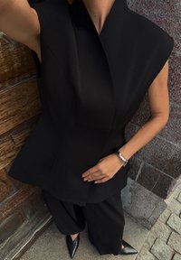 Zwart getailleerde top met een hoge kraag en brede mouwen, gecombineerd met losse zwarte broeken. De outfit heeft een zachte stof en scherpe hoeken.