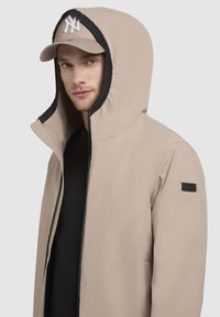 Giacca a vento beige con fodera interna nera e patch del marchio. Indossata sopra una maglietta nera, abbinata con un cappellino beige con logo bianco.