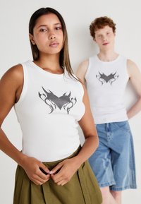 Balti, banguoti, be rankovių marškiniai su juodu ugnies grafiku ant krūtinės, dėvimi su žalia plisuota sijonu ir džinsiniais šortais.