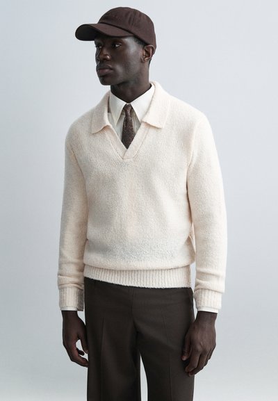 Pull rose clair en maille avec un col en V et un ourlet côtelé, associé à une chemise blanche à col, une cravate marron à motifs, une casquette marron et un pantalon foncé.