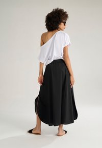 Top bianco a spalle scoperte abbinato a una gonna nera lungo la caviglia con spacco laterale. La modella indossa sandali neri. Tessuto morbido e casual.