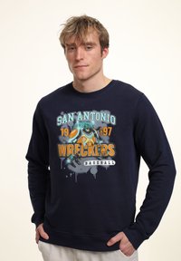 Marinblå sweatshirt med grafisk design som har texten "San Antonio Wreckers Baseball", föreställande en robot med orange och blå detaljer, rund halsringning.