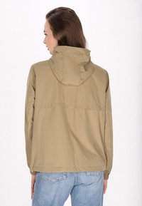 Giacca cappuccio beige con una texture leggera e liscia. Presenta polsini elasticizzati, un dettaglio sul retro e una vestibilità rilassata sopra un paio di jeans blu.