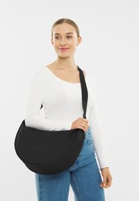 Czarna torba na ramię z tkaniny o zakrzywionym kształcie, regulowanym pasku, gładkiej fakturze i subtelnym wytłoczonym logo z przodu.