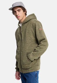 Olijfgroene hoodie met rits en een gestructureerde brei-afwerking, met een kangoeroezak, een trekkoordhoed en een leren logo-patch op de mouw.