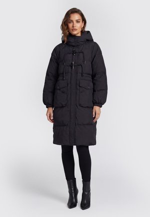 Frau trägt einen langen schwarzen Steppmantel mit Kapuze, Vordertaschen mit Klappen, Knebelverschlüsse, schwarze Leggings und schwarze Stiefeletten.