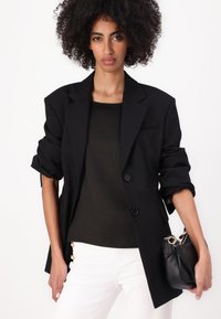 Blazer nero oversize con maniche arrotolate, chiusura con un solo bottone, abbinato a un top scuro e pantaloni bianchi con dettagli in perle. Borsa nera.