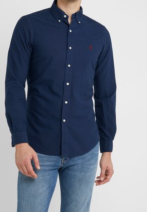 Mand iført en mørkeblå skjorte med knapper og lange ærmer og lyseblå jeans, stående mod en ensfarvet lys grå baggrund.