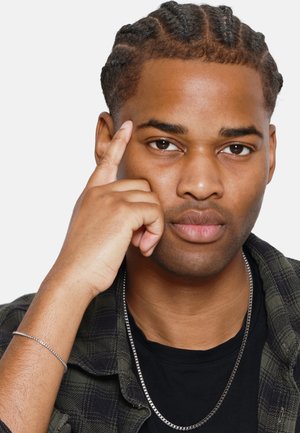 Portret van een mannelijke modellen met gevlochten donker haar, gekleed in een zwart shirt en een groen geruite jas, met zilveren kettingen en armbandaccessoires.