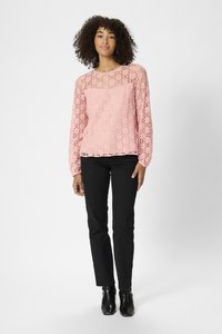 Haut en dentelle rose clair avec des manches longues et un col festonné, associé à un pantalon noir et des bottines. Détails de motif en dentelle complexe visibles.